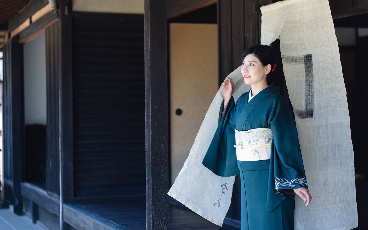 暖簾のかかる町家の前に立つ深緑の正絹の着物姿の女性